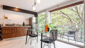 A modern kitchen with a glass dining table, black chairs, and a view of trees through large windows. - w* | Enchanting 1BR w/ Terrace in Prime Polanco (Ciudad de México)