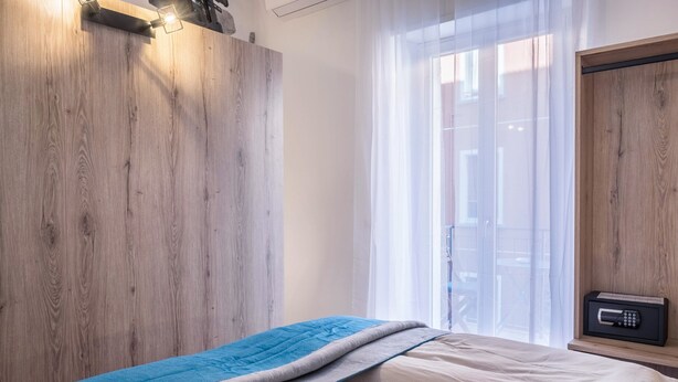 A bedroom with a wooden wardrobe, a bed with beige and blue bedding, a window with sheer curtains, and a radio on a shelf. - Apartment "Isabel" with Mountain View, French Balcony & Wi-Fi (Bolzano)