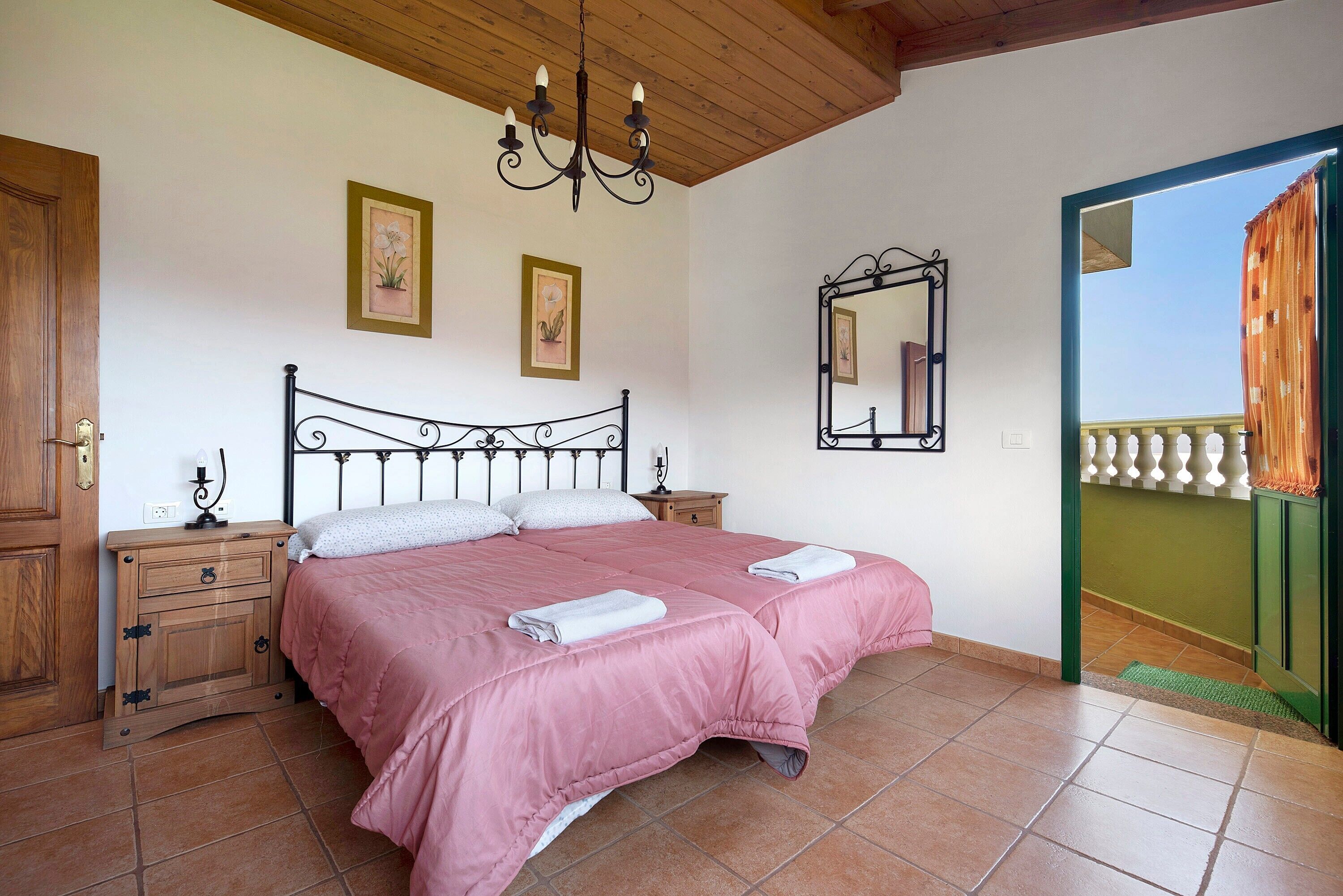 Un dormitorio con cama, mesitas de noche de madera, candelabro, espejo y vista al balcón.