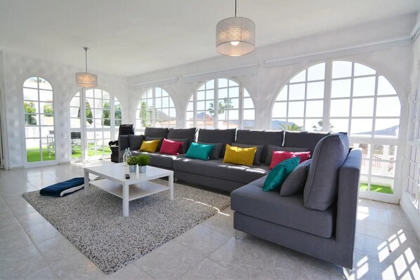 Ein geräumiges Wohnzimmer mit grauem Sofa, farbenfrohen Kissen, einem weißen Couchtisch und großen Bogenfenstern.