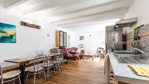 A dining area with a wooden table and chairs, a kitchen with a sink and stove, and a living area with a sofa and a painting on the wall. - Vacation Home "Gite Senguinous 1" with Shared Pool, Private Terrace & Wi-Fi (Lesparre-Médoc)
