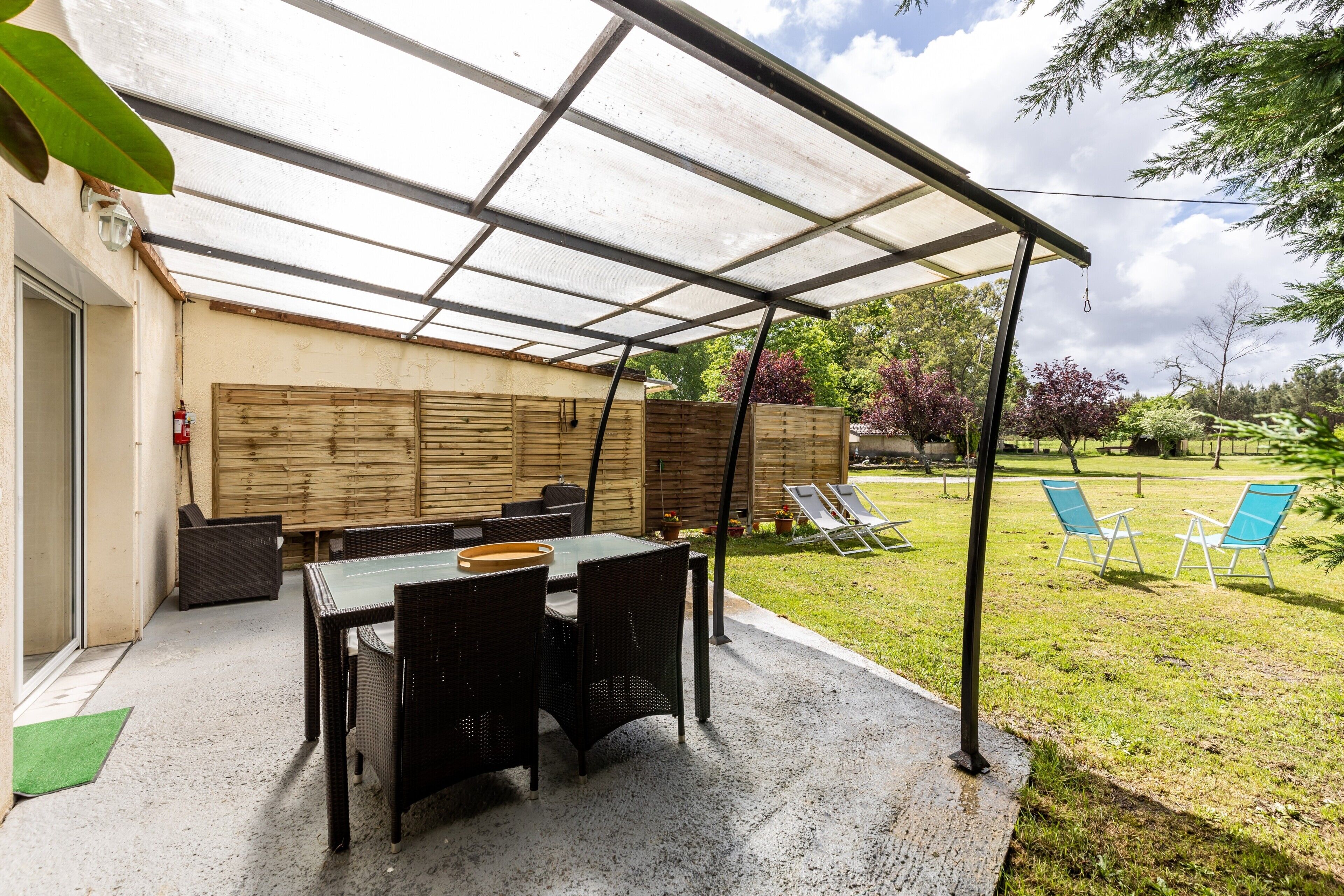 Een terras buiten met een tafel en stoelen, een overdekt gedeelte en een gazon met zitmogelijkheden.