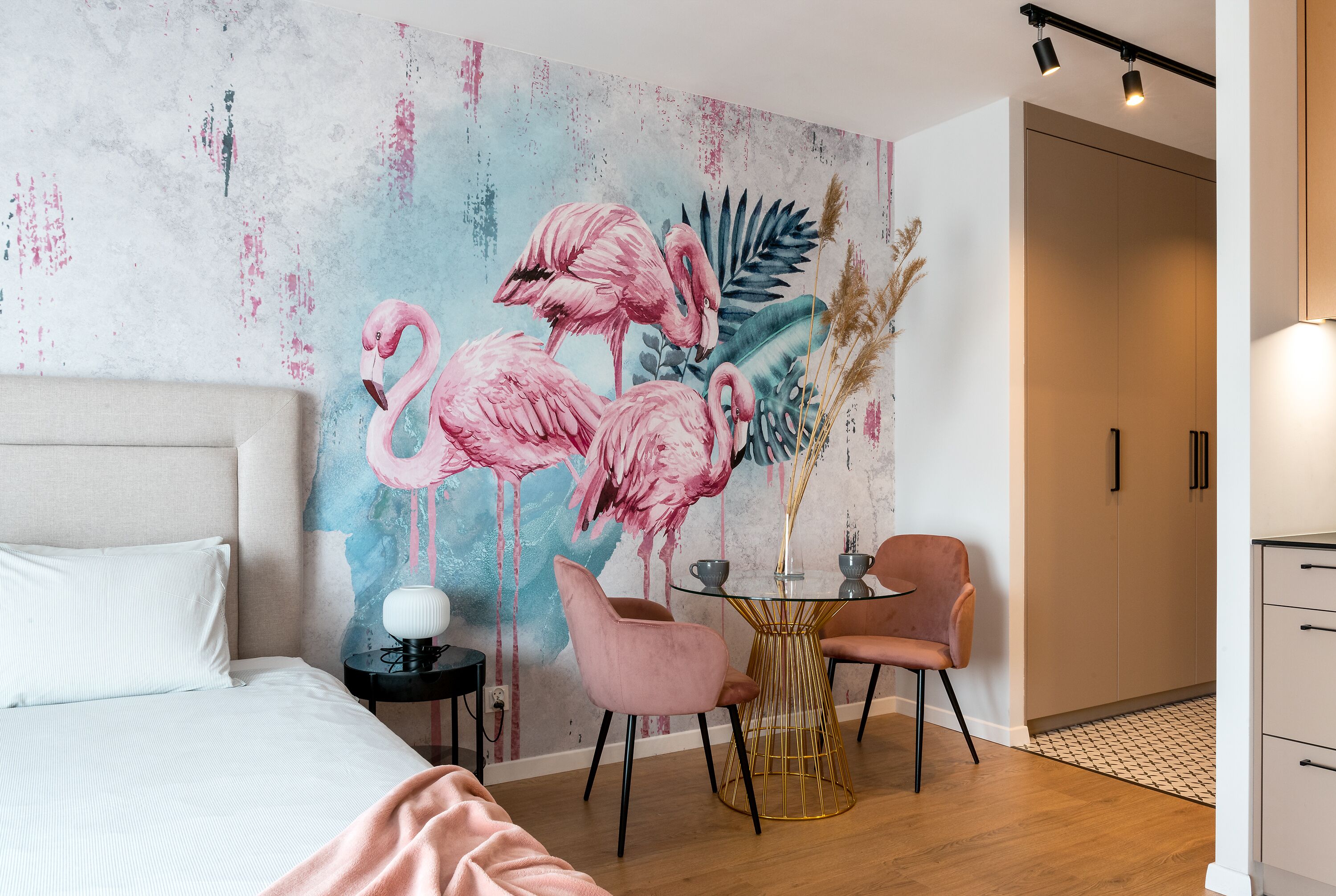 A bedroom with a bed, a nightstand, a table with chairs, and a wall mural of flamingos.