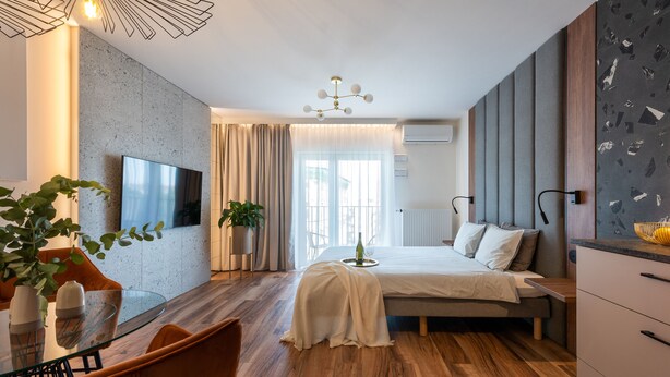 Habitació d'hotel moderna amb taula de dinar de vidre, llit amb capçalera gris, finestra gran amb cortines i televisió penjada a la paret.