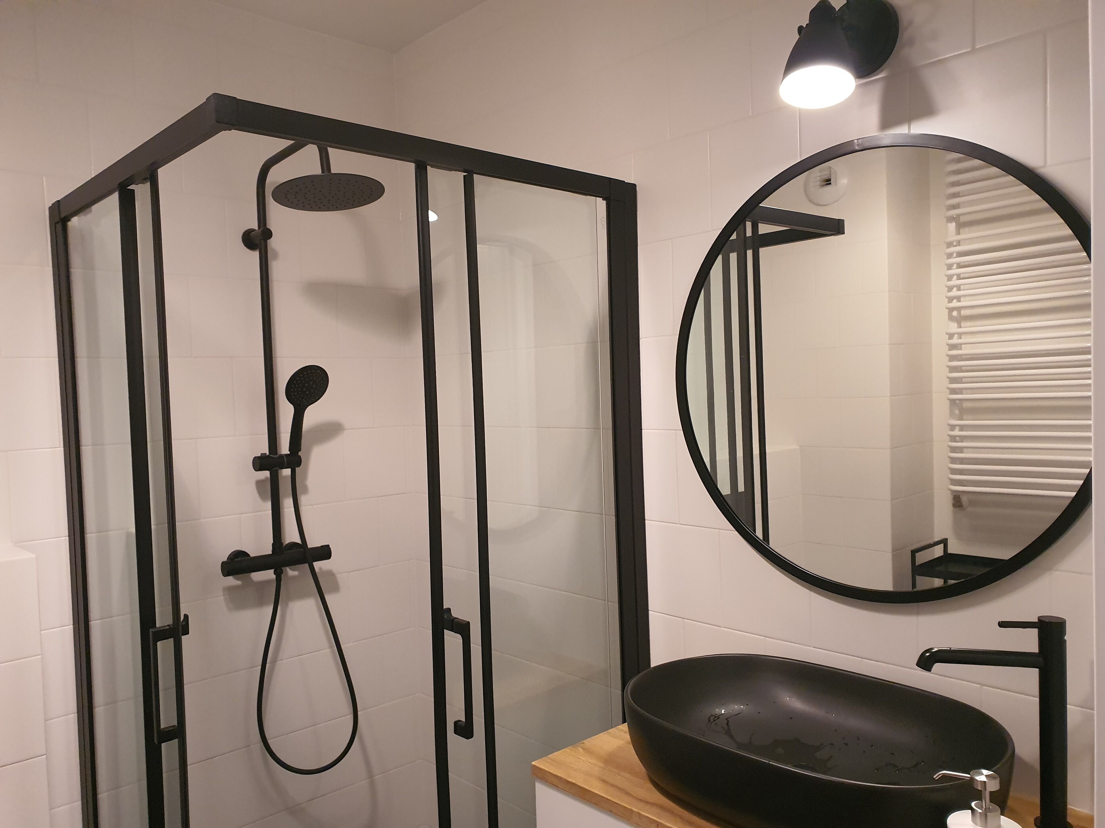 A modern bathroom with a glass-enclosed shower, a round mirror, and a black sink on a wooden countertop.