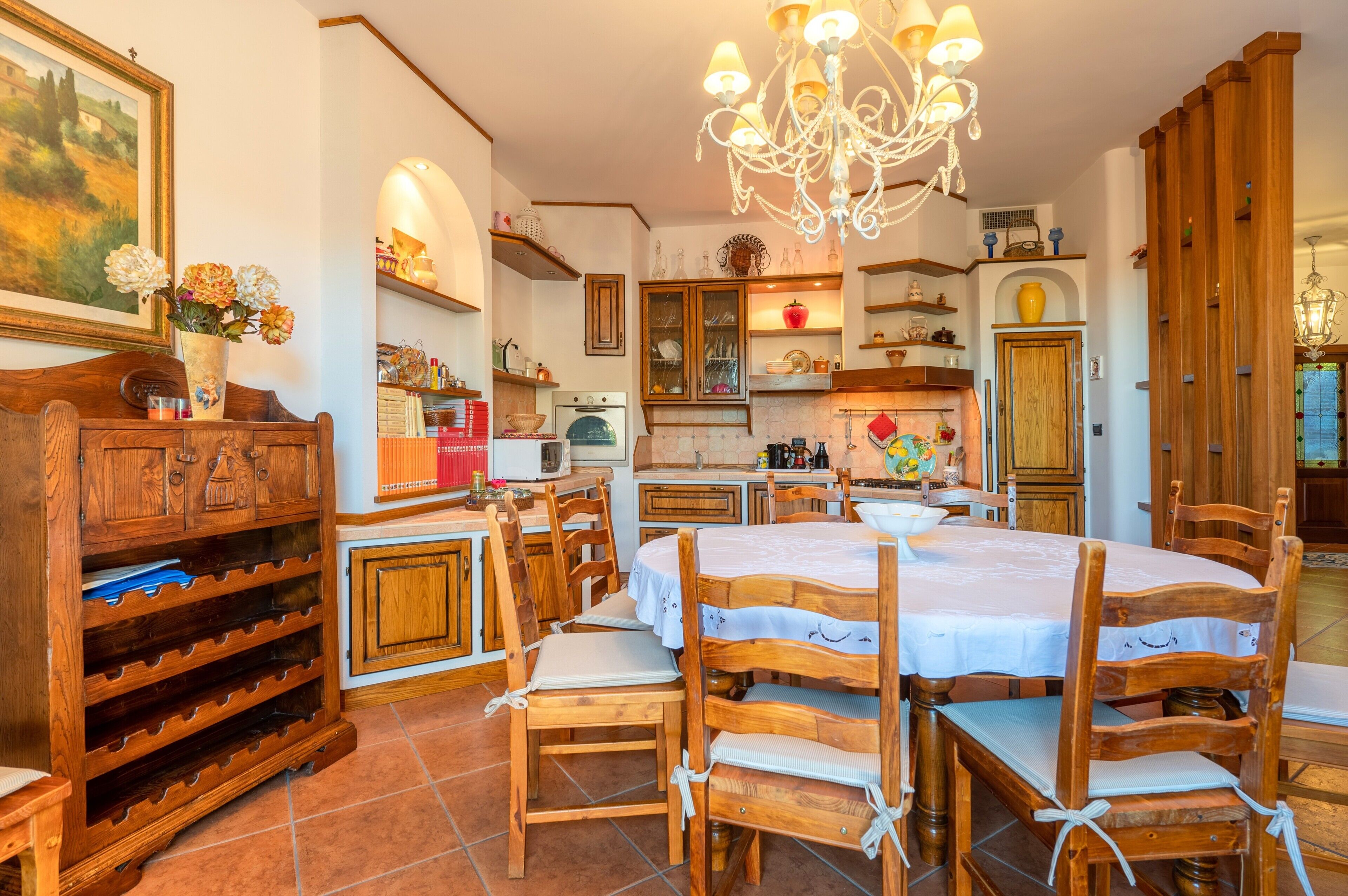 A dining area with wooden chairs and a table, a chandelier, and a kitchen in the background.