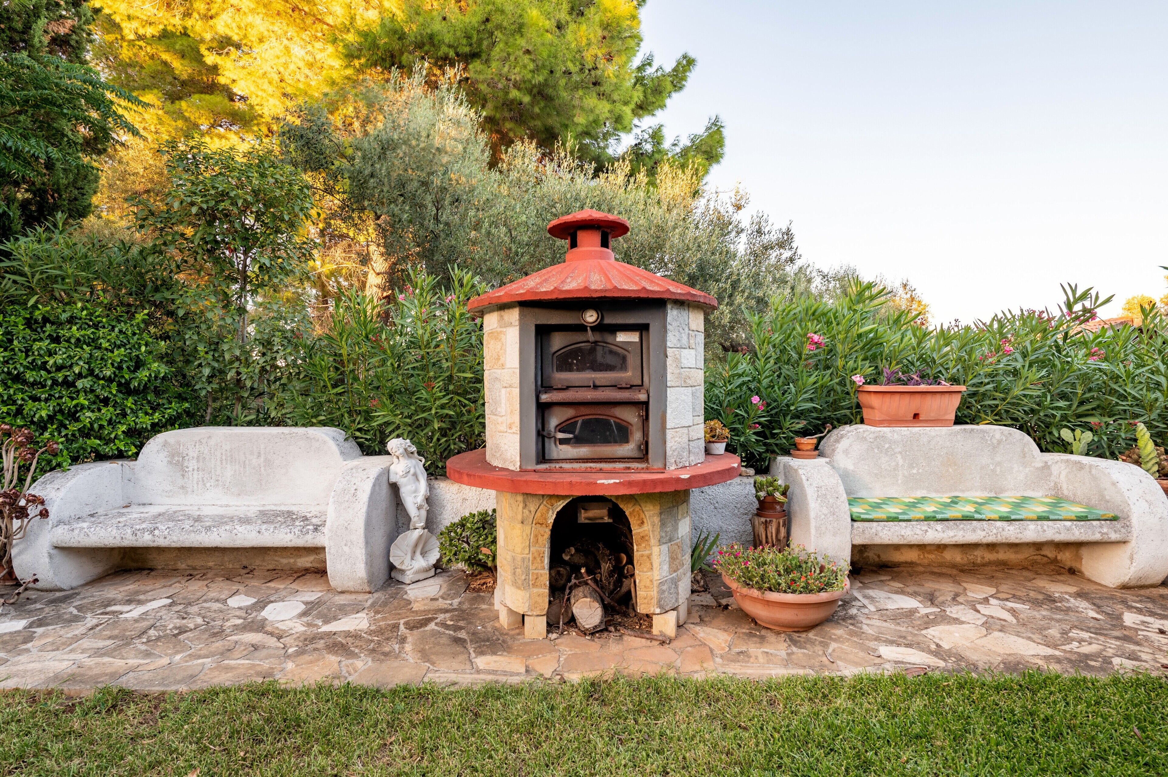 Un área de estar al aire libre de piedra con un horno de leña incorporado, rodeada de plantas en macetas y un césped delantero.