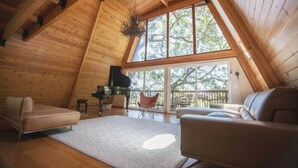 A cozy attic room with wooden walls, a piano, a sofa, and a large window with a view of trees. - Luxurious Bayview Treehouse: Hot Tub, 15 min to SF (Brisbane)