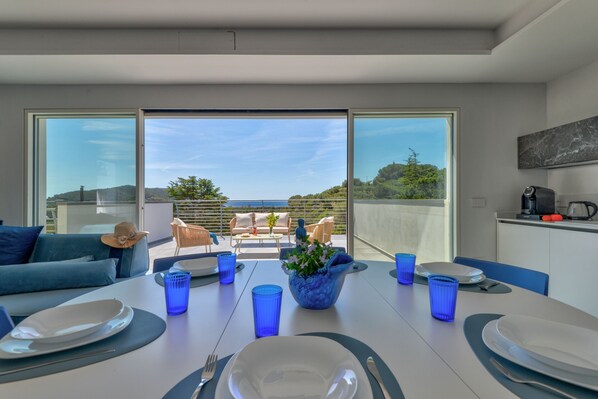 Ein moderner Essbereich mit Tisch für vier Personen, große Fenster mit Blick auf einen Hügel und eine Küchenzeile mit Kaffeemaschine und Toaster.