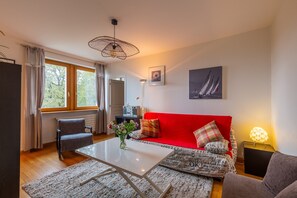 A living room with a red sofa, a white coffee table, a grey armchair, a patterned rug, and a painting of sailboats on the wall. - Apartment '2p au Belvédère' with Lake view and Balcony (Thonon-les-Bains)