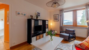 Una sala de estar con una mesa de centro blanca, un mueble de entretenimiento negro, un sillón gris, un sofá con un cojín rojo y vistas a la ciudad a través de una ventana.