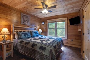 Une chambre de style cabane en bois, avec un lit, deux lampes de chevet, un ventilateur de plafond, une horloge, une fenêtre et un téléviseur fixé au mur.