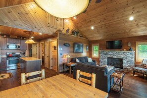 A cozy cabin interior with a kitchen, dining area, and living room featuring wooden floors and a stone fireplace.