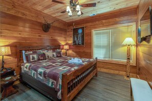 A wooden cabin bedroom with a bed, a ceiling fan, a window with blinds, and a wall-mounted lamp.