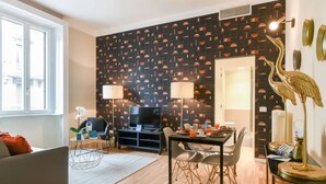 Ein modernes Wohnzimmer mit einer Wand in Schwarz-Weiß-Muster, einem Flachbildfernseher, einem Esstisch mit Stühlen und einer dekorativen, goldenen Vogelstatue.