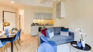 Un espace de vie compact comprenant un coin repas, une kitchenette et un canapé gris agrémenté de coussins bleus.