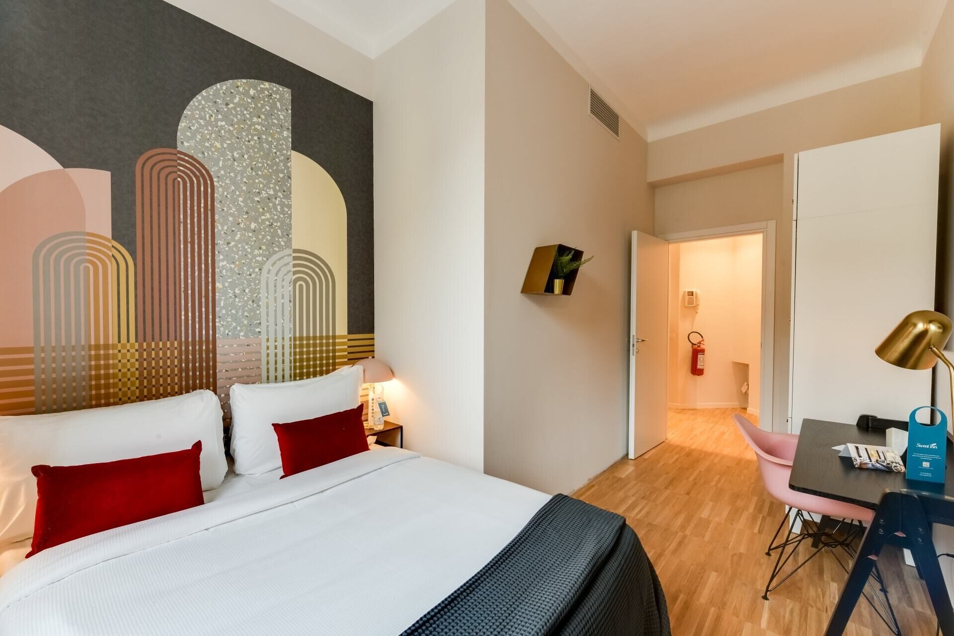 Habitación de hotel con cama, escritorio, silla, una obra de arte en la pared y una puerta que da a otra habitación.