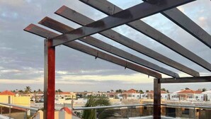 Una terraza en la azotea con una pérgola, dos sillas de mimbre blanco y una mesita con dos jarrones amarillos.
