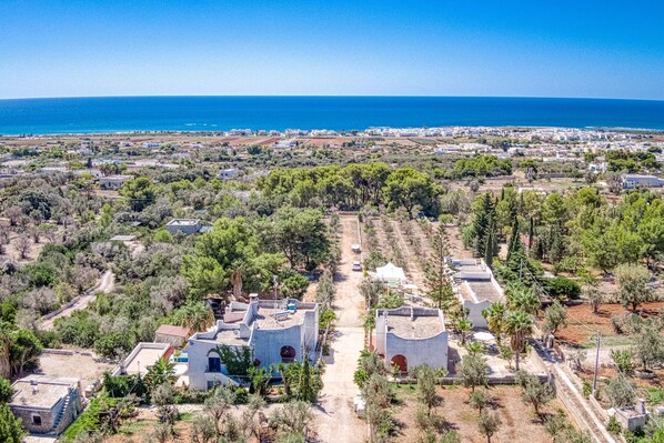 An aerial view of a coastal town with a clear road leading to a cluster of buildings, surrounded by greenery and the sea in the background.