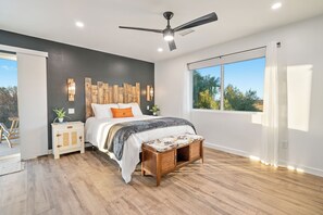 A bedroom with a bed, bedside tables, a ceiling fan, a window with a view, and a wooden floor.