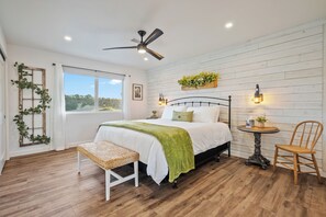 Quarto com cama, um banquinho, mesa, cadeira, ventilador de teto, janela e um vaso fixado na parede.