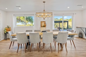 A dining area with a wooden table, six grey upholstered chairs, a chandelier, and large windows offering a view of trees.