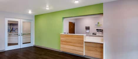 A reception area with a wooden reception desk, a green accent wall, and a glass door leading to a room with a visible bed.