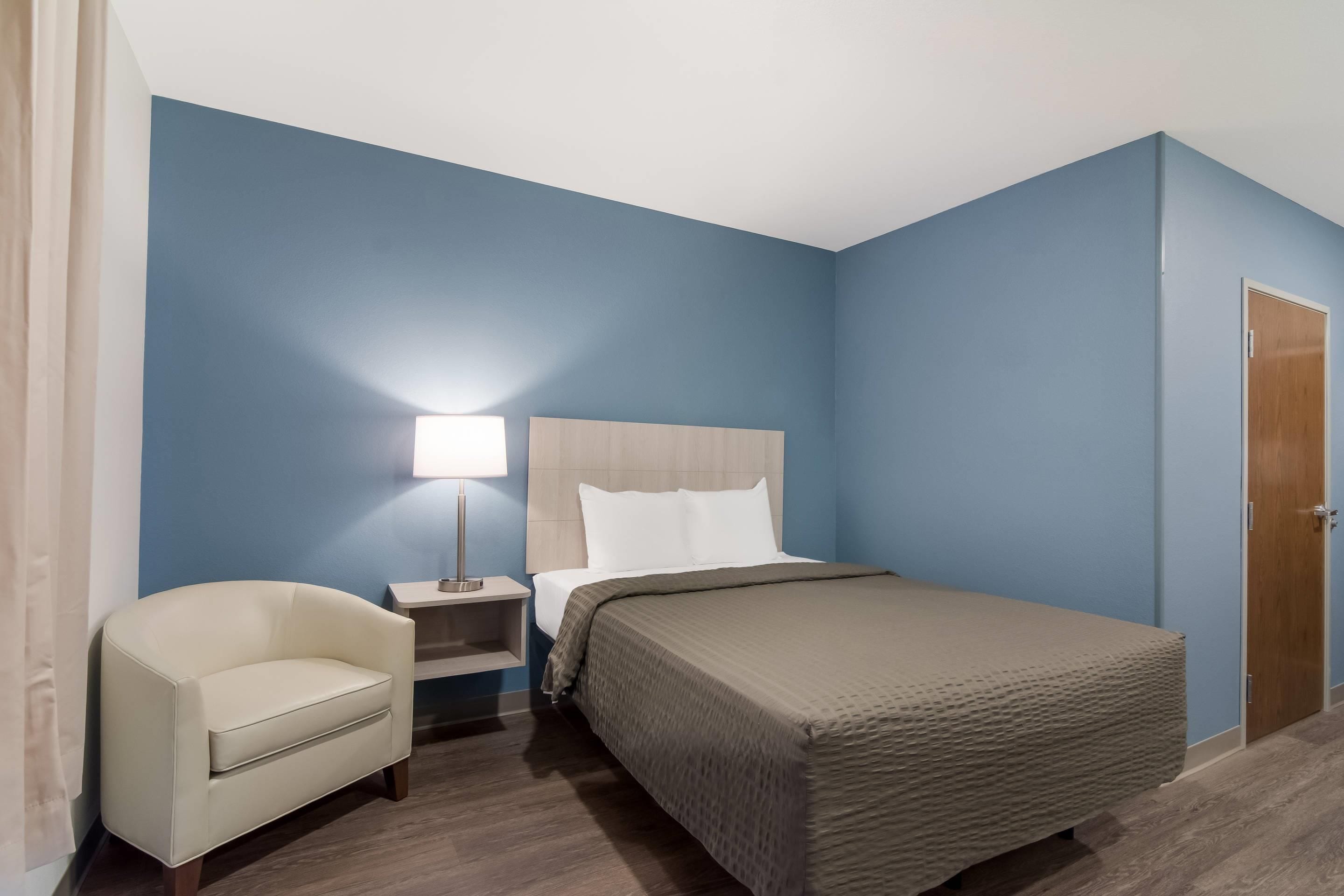 Una habitación de hotel con cama, mesita de noche, lámpara, silla y puerta.