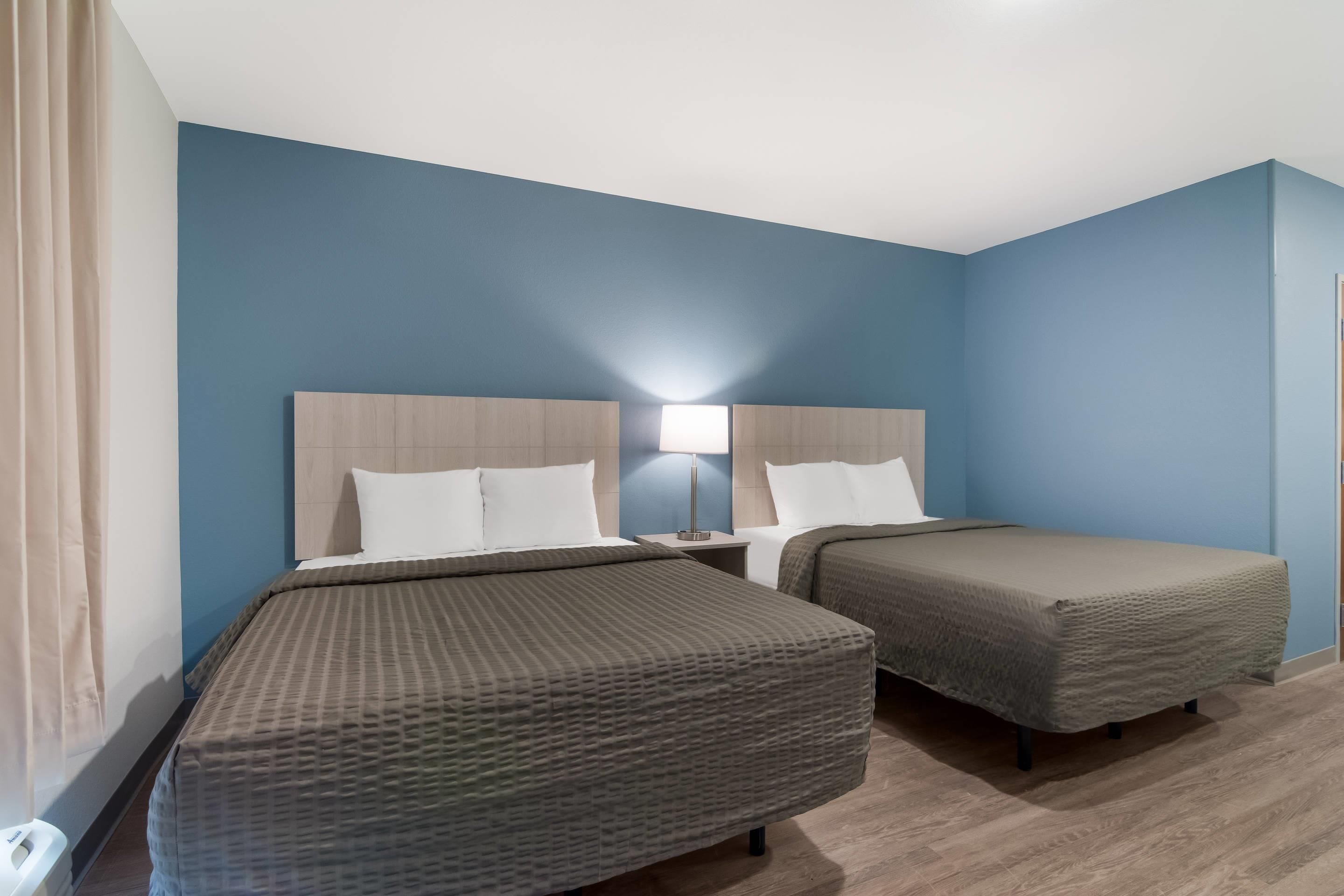 A hotel room with two beds, a nightstand, a lamp, and a window with curtains.