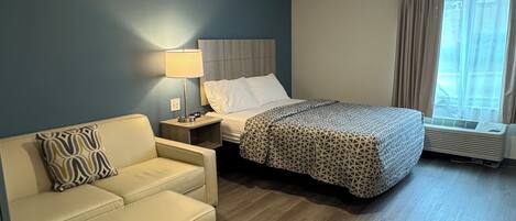 Habitación de hotel con cama, sofá, mesita de noche, lámpara y ventana con cortinas.