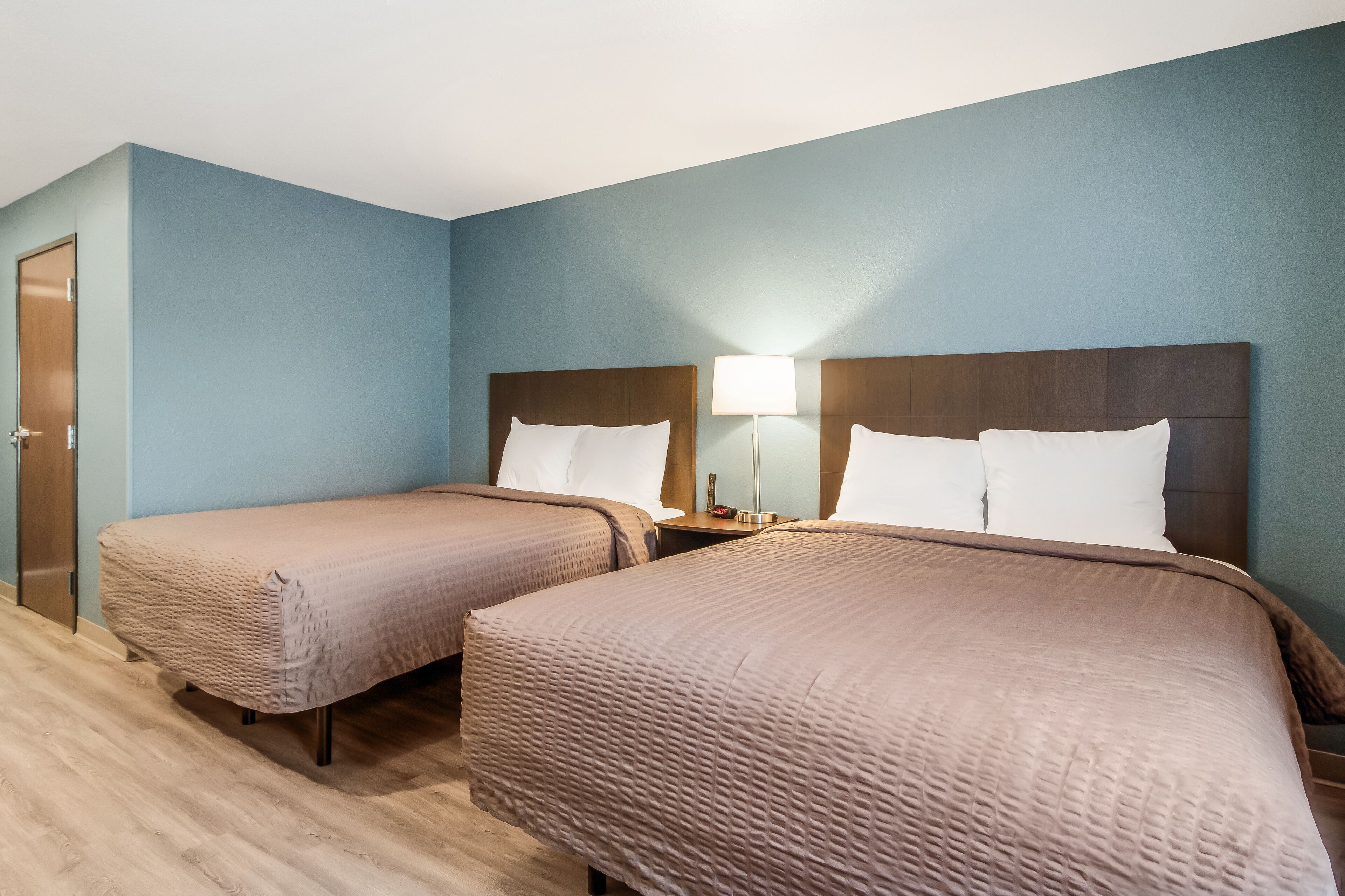 A hotel room with two single beds, a wooden door, and a nightstand with a lamp.