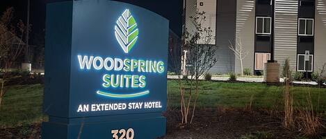 A sign for Woodspring Suites, an extended stay hotel, with a modern building in the background.