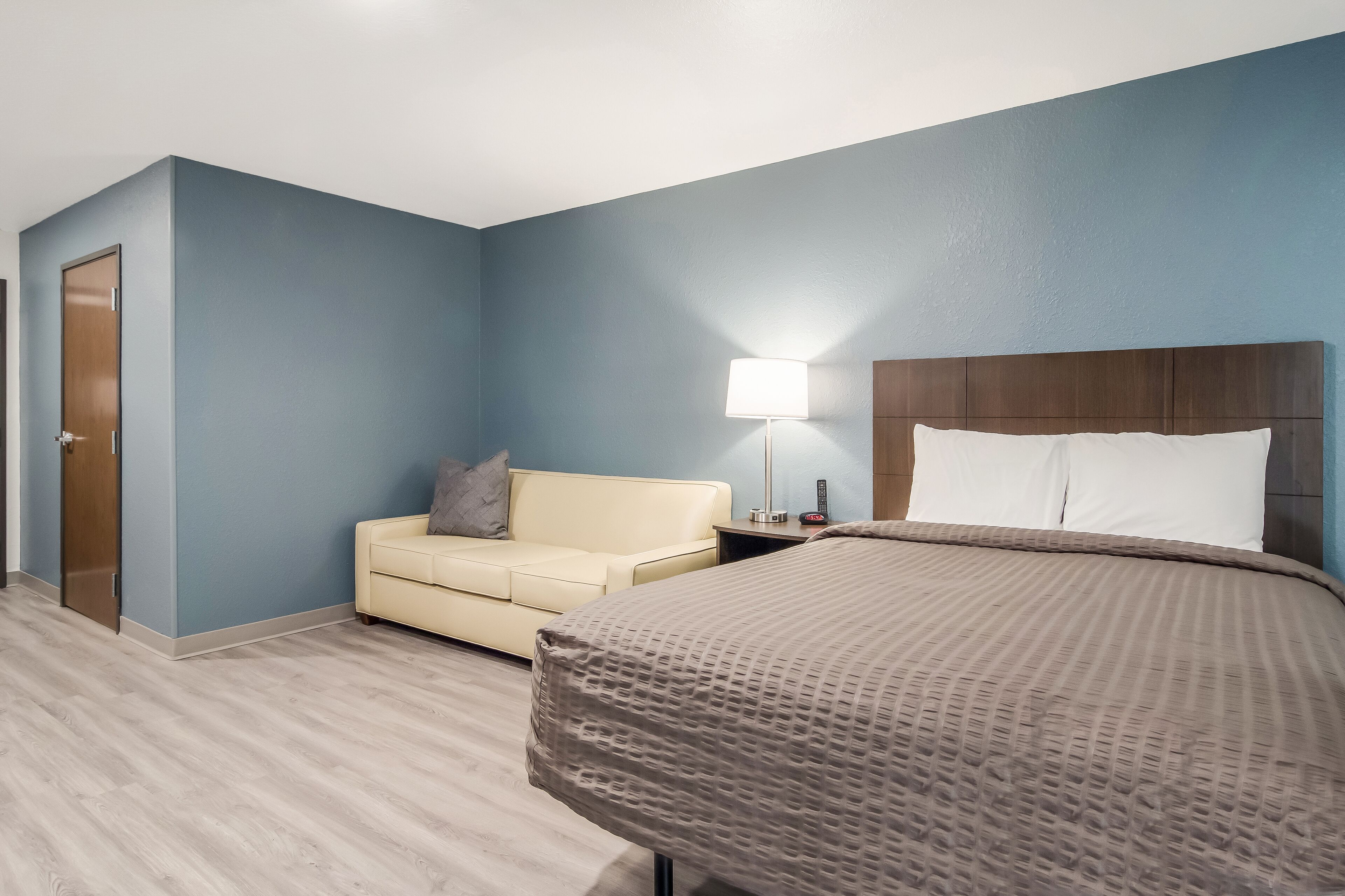 A hotel room with a bed, a bedside table, a lamp, a sofa, and a door.