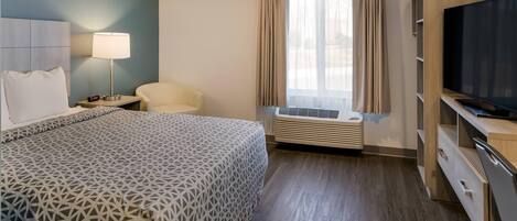 Quarto de hotel com cama, poltrona, televisão e janela com cortinas.