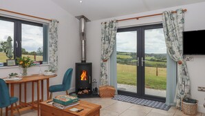 A cozy living room with a fireplace, a dining area, and large windows offering a view of the countryside. - The Pennymoor (Tiverton)