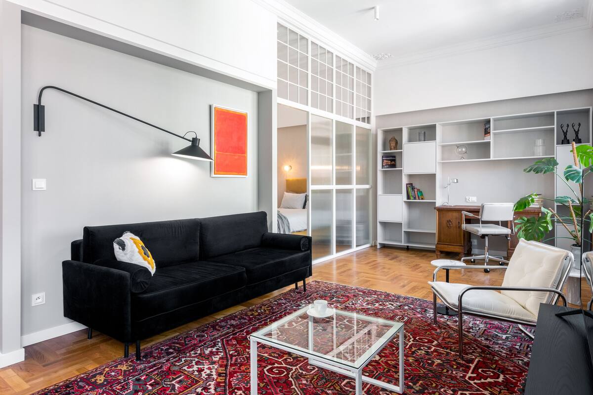 A modern living room with a black sofa, a glass coffee table, and a red patterned rug.