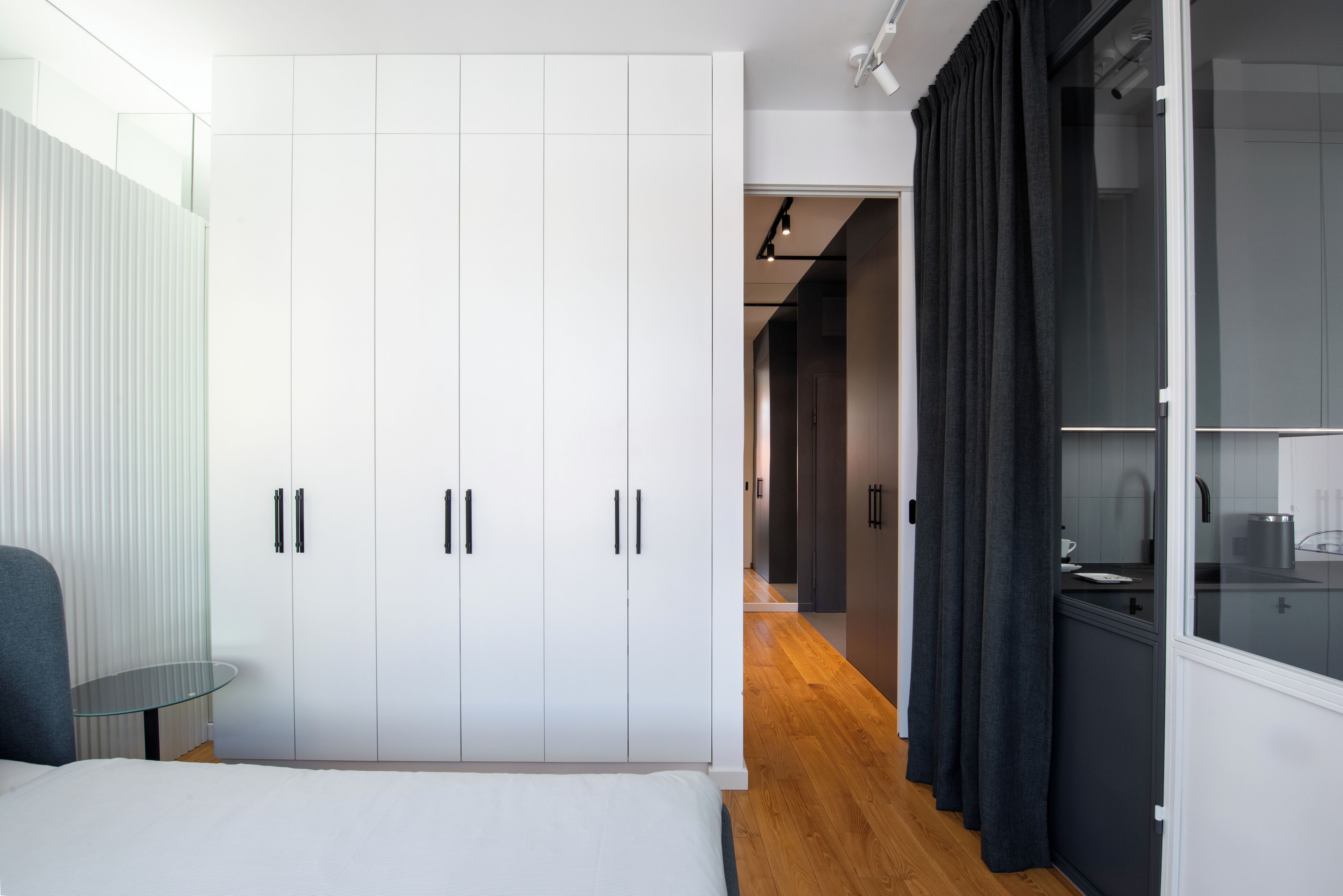 A bedroom with a bed, a glass table, white wardrobes, a dark kitchen area, and a wooden floor.