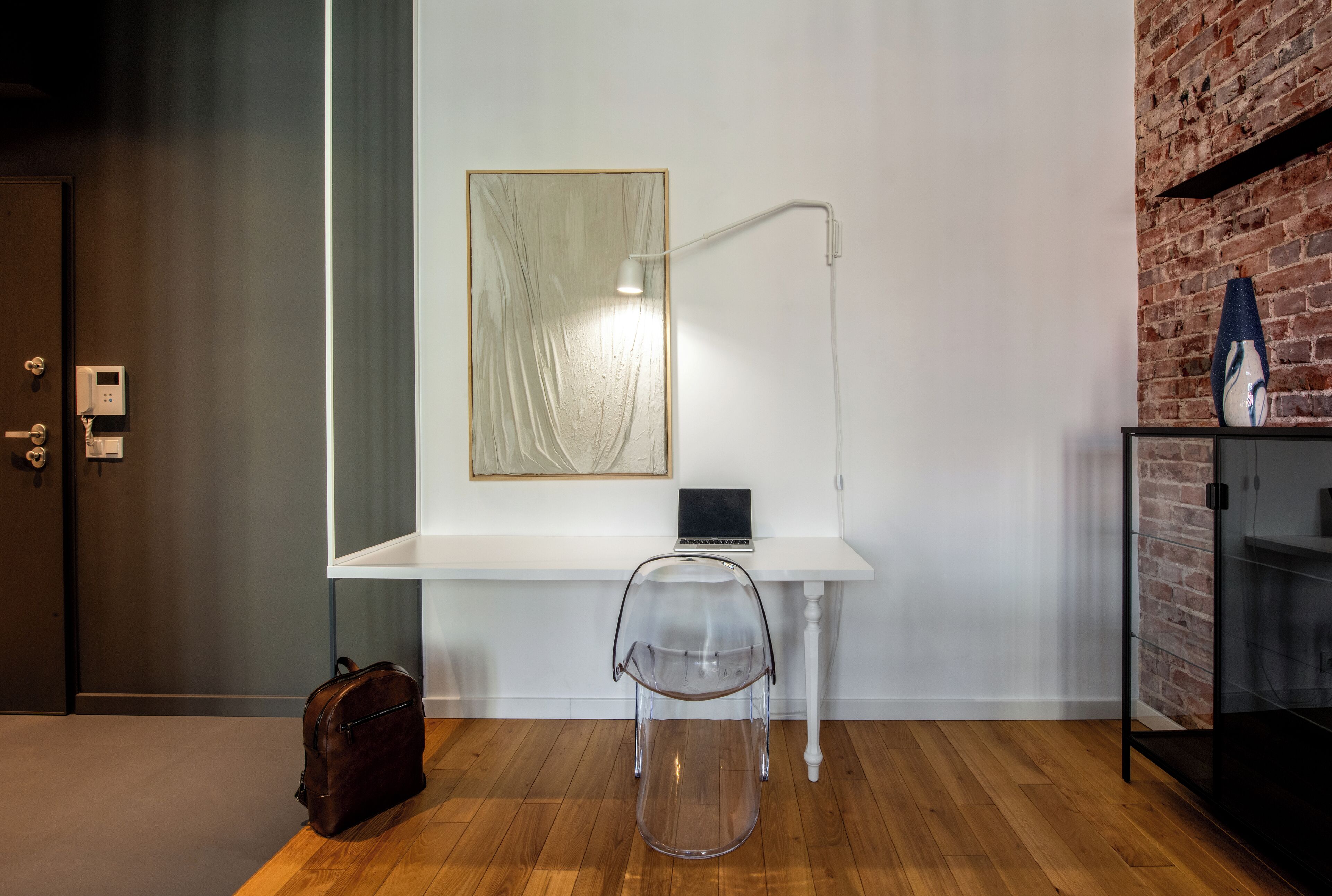 Una habitación moderna con un escritorio blanco, una silla transparente, una computadora portátil, una lámpara, una pared de ladrillo y suelo de madera.