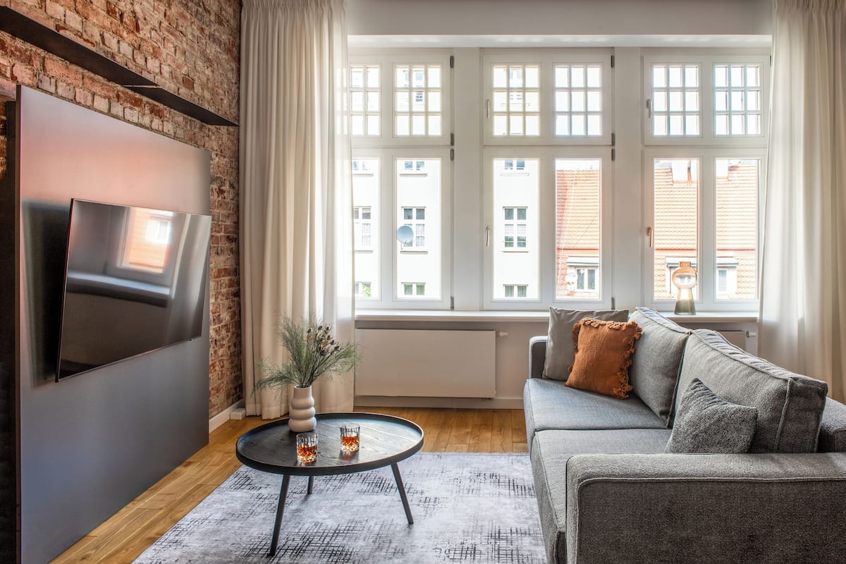 A modern living room with a flat-screen TV mounted on a brick wall, a grey sofa, a round coffee table with decorative items, and large windows with curtains.