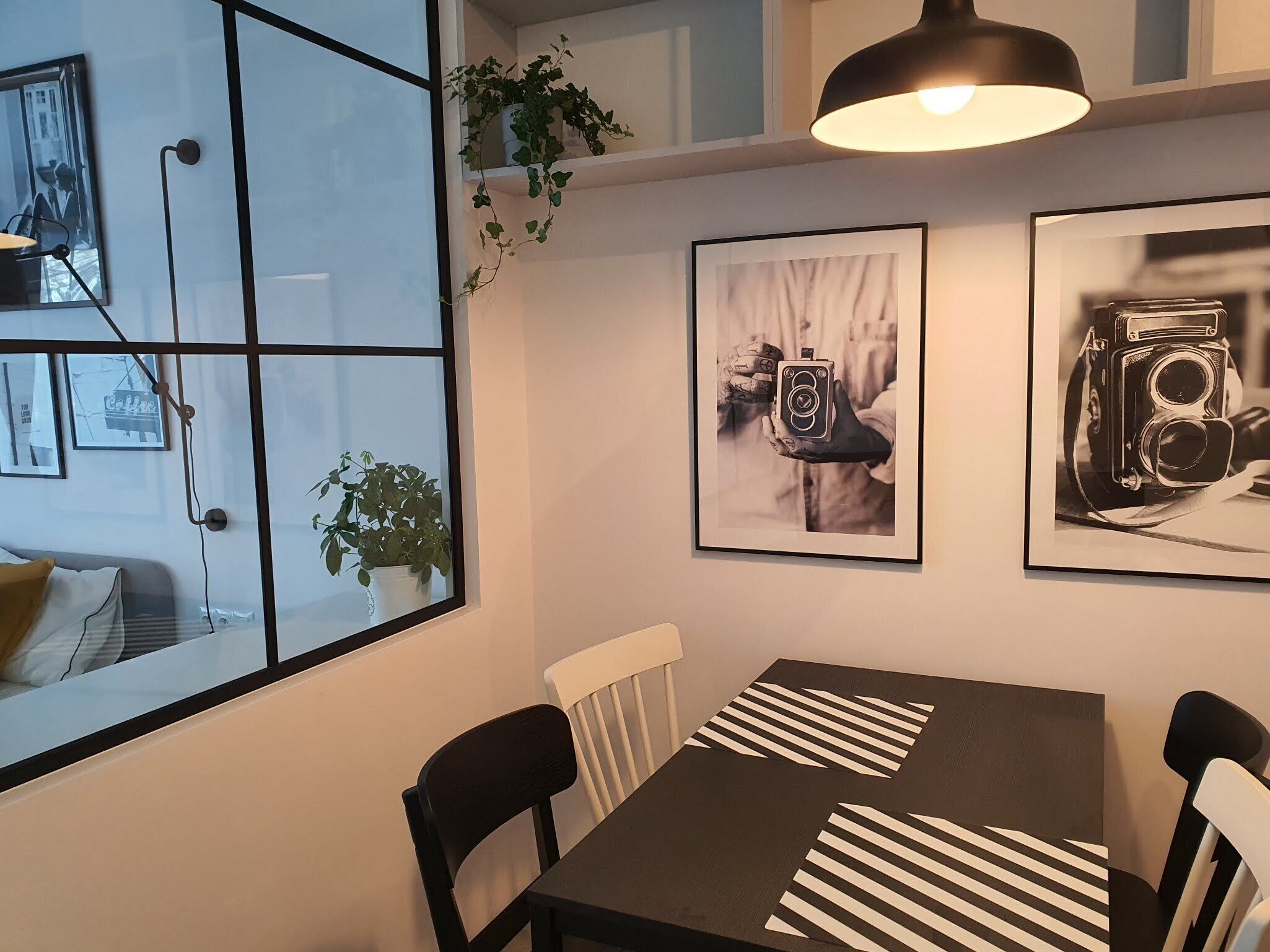 Et moderne spiseområde med et mørkt træbord, stribede servietter og sorte stole. Væggen er prydet med indrammede fotografier, og der hænger en plante samt en pendellampe over bordet.