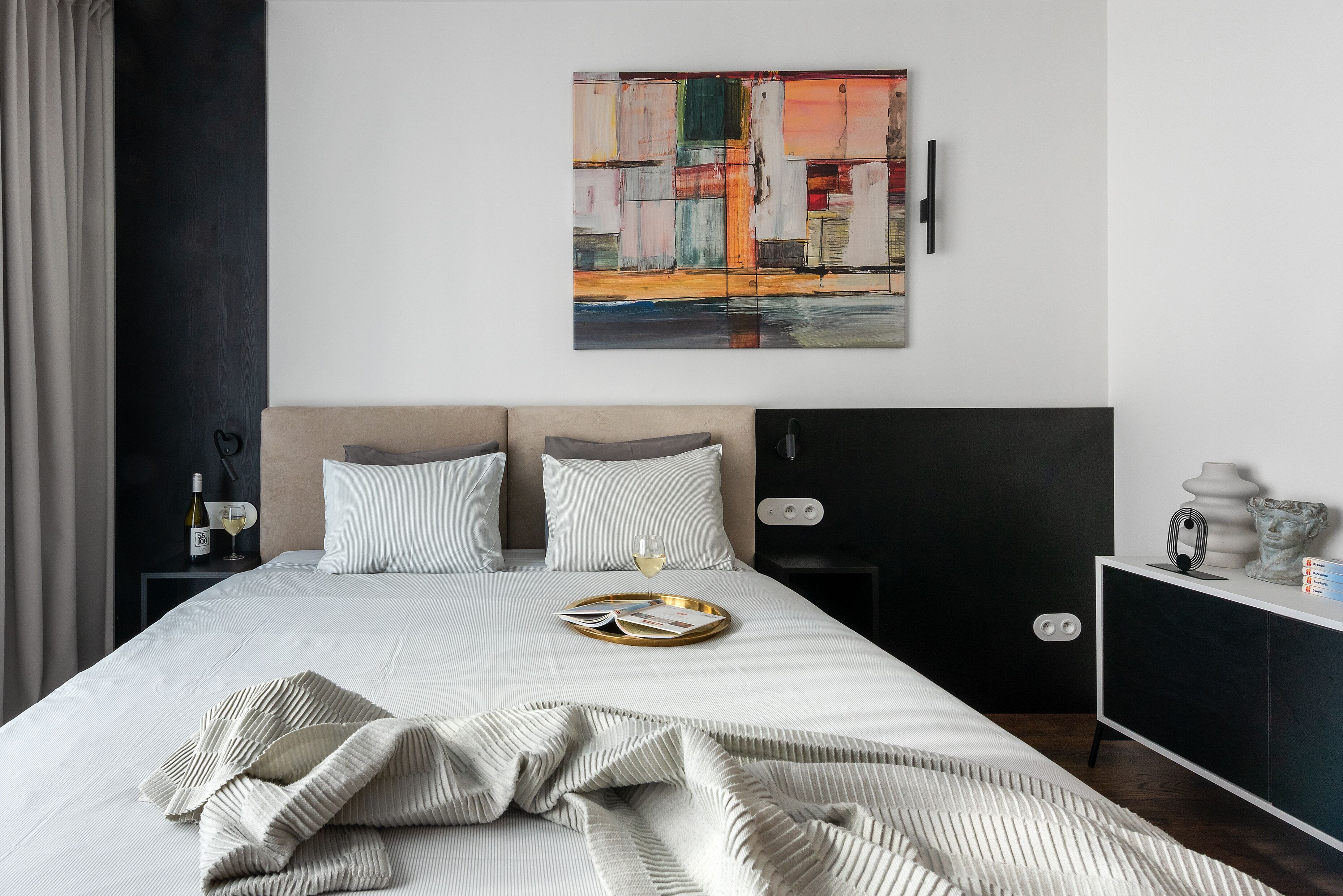 A neatly made bed with a tray of magazines and a glass of wine, a modern abstract painting on the wall, and a minimalist nightstand with a lamp.