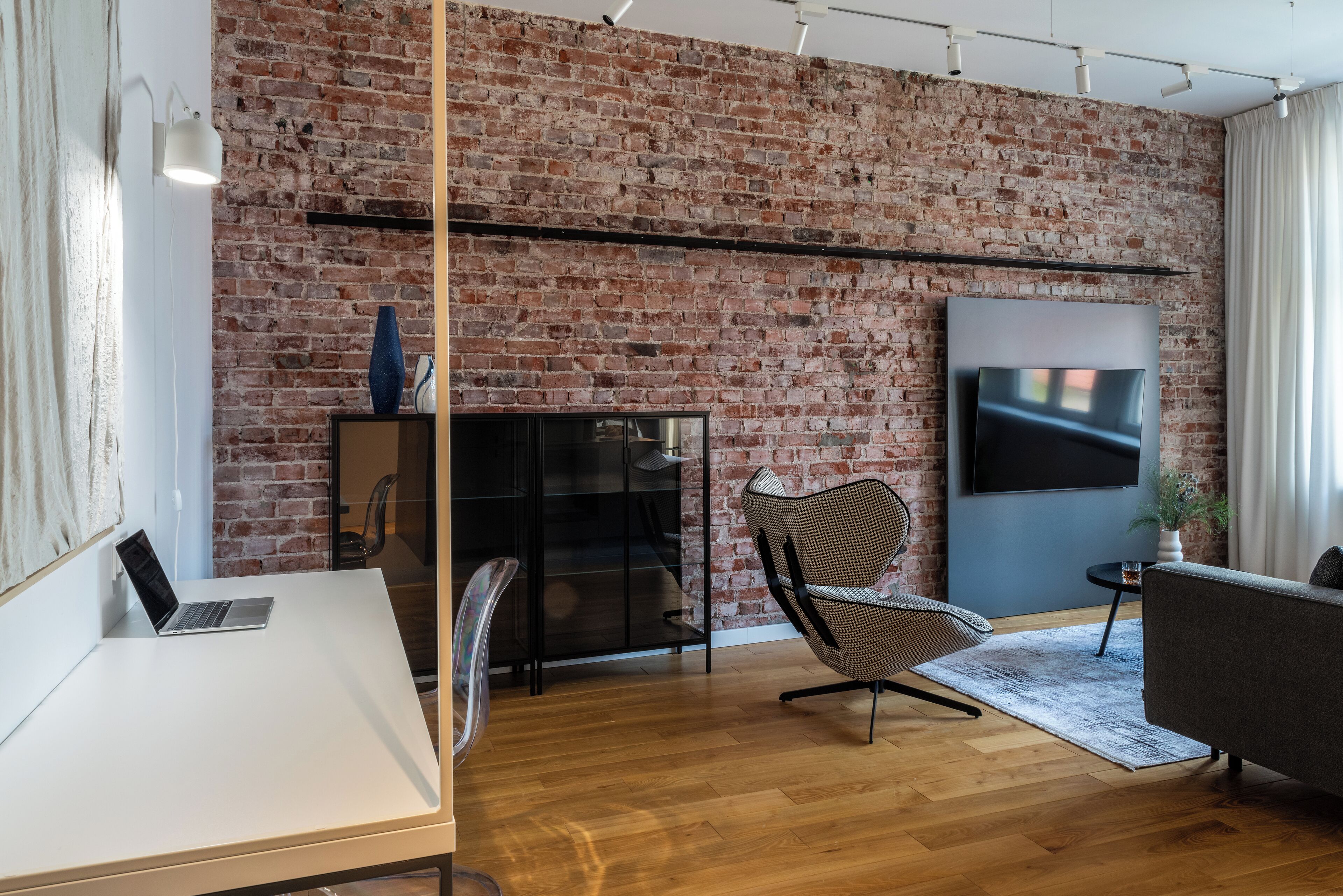 A modern living room with a brick wall, a flat-screen TV, a wooden floor, a white desk, and a black chair.