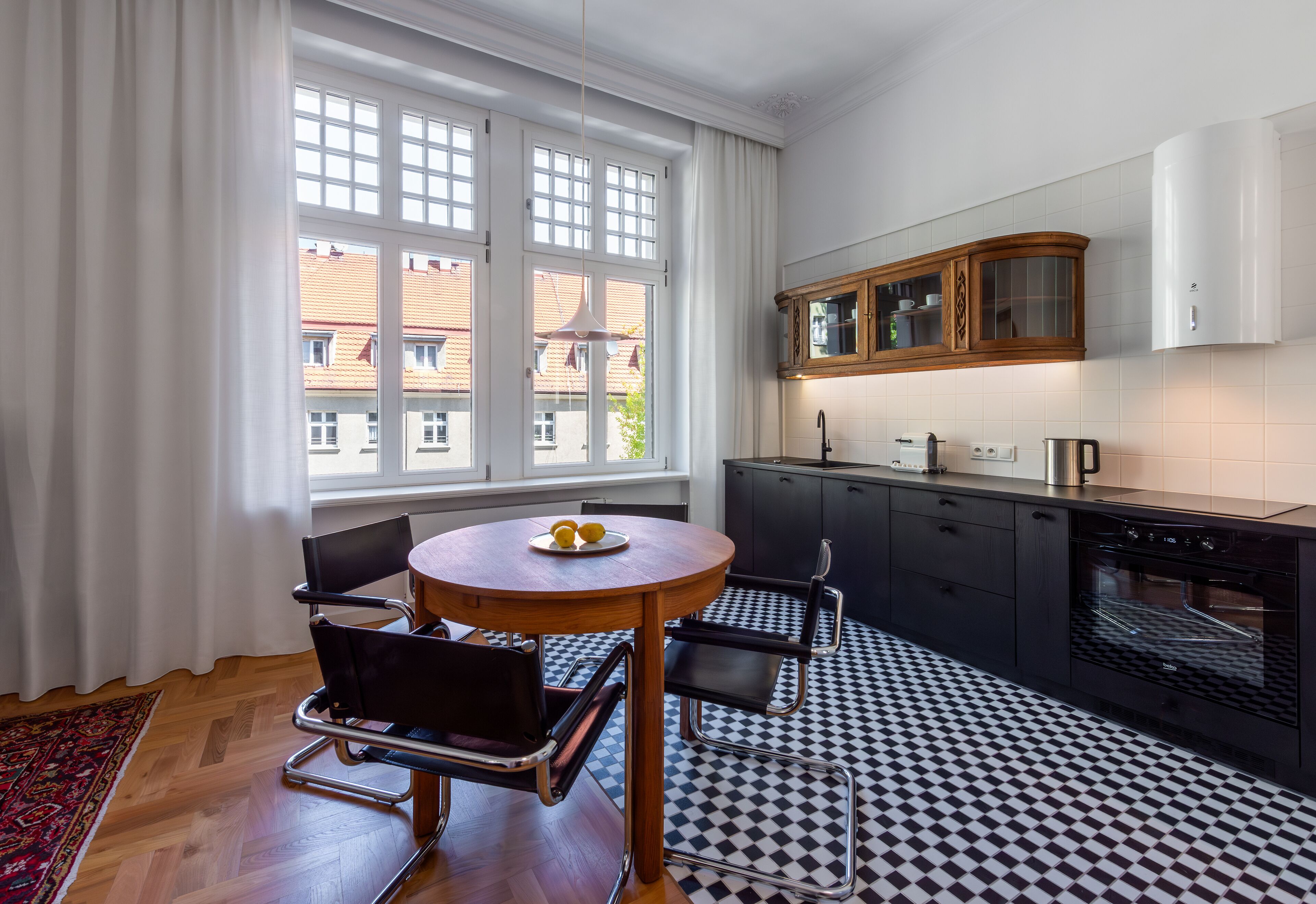 Une cuisine moderne avec un sol à carreaux noirs et blancs, une table ronde en bois accompagnée de deux chaises, et une fenêtre ornée de rideaux blancs.