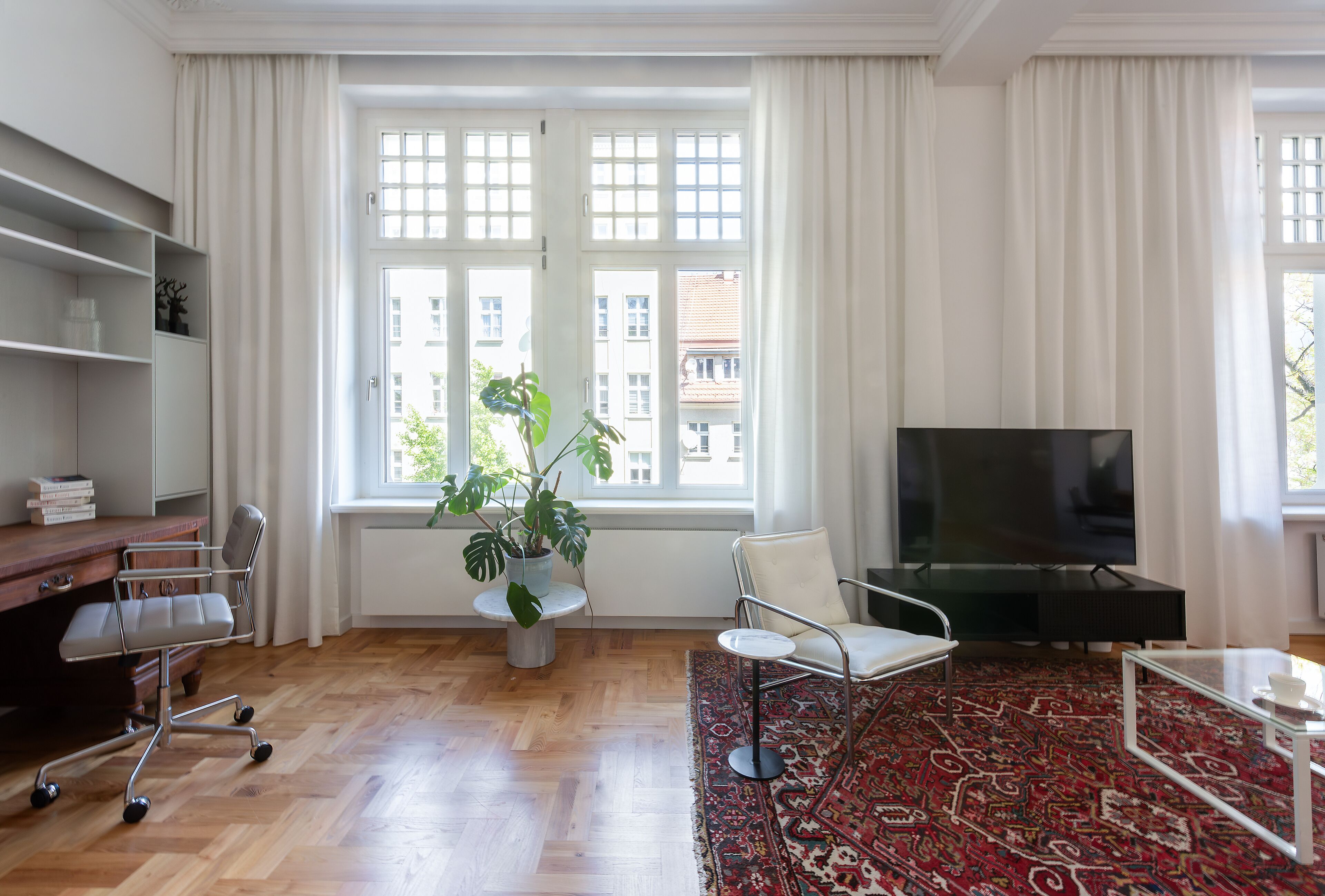 A modern living room with a wooden floor, a large TV, a white chair, a desk with a chair, a potted plant, and a patterned rug.