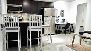 Una cocina moderna con armarios oscuros, microondas y refrigerador. Hay sillas blancas y una mesa de comedor con un florero.