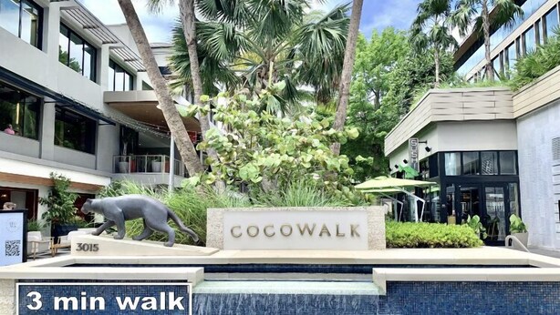Señalización de 'Cocowalk' a 3 minutos a pie, rodeada de palmeras y un elemento acuático.