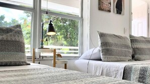 Un dormitorio con un ventanal grande, una cama con ropa de cama gris, una mesita de noche con lámpara y una obra de arte en la pared.