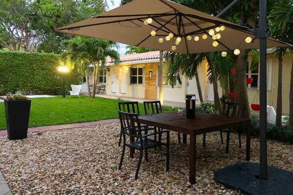 An outdoor patio with a table and chairs under a canopy, surrounded by a gravel area and greenery.
