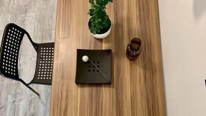 Un tavolo da pranzo in legno con due sedie nere, una pianta in vaso, un piccolo oggetto nero e un bicchiere con un liquido.