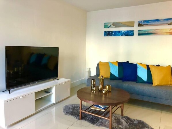 Ein modernes Wohnzimmer mit einem Flachbildfernseher auf einem weißen Schrank, einem grauen Sofa mit bunten Kissen und einem Couchtisch mit dekorativen Accessoires.